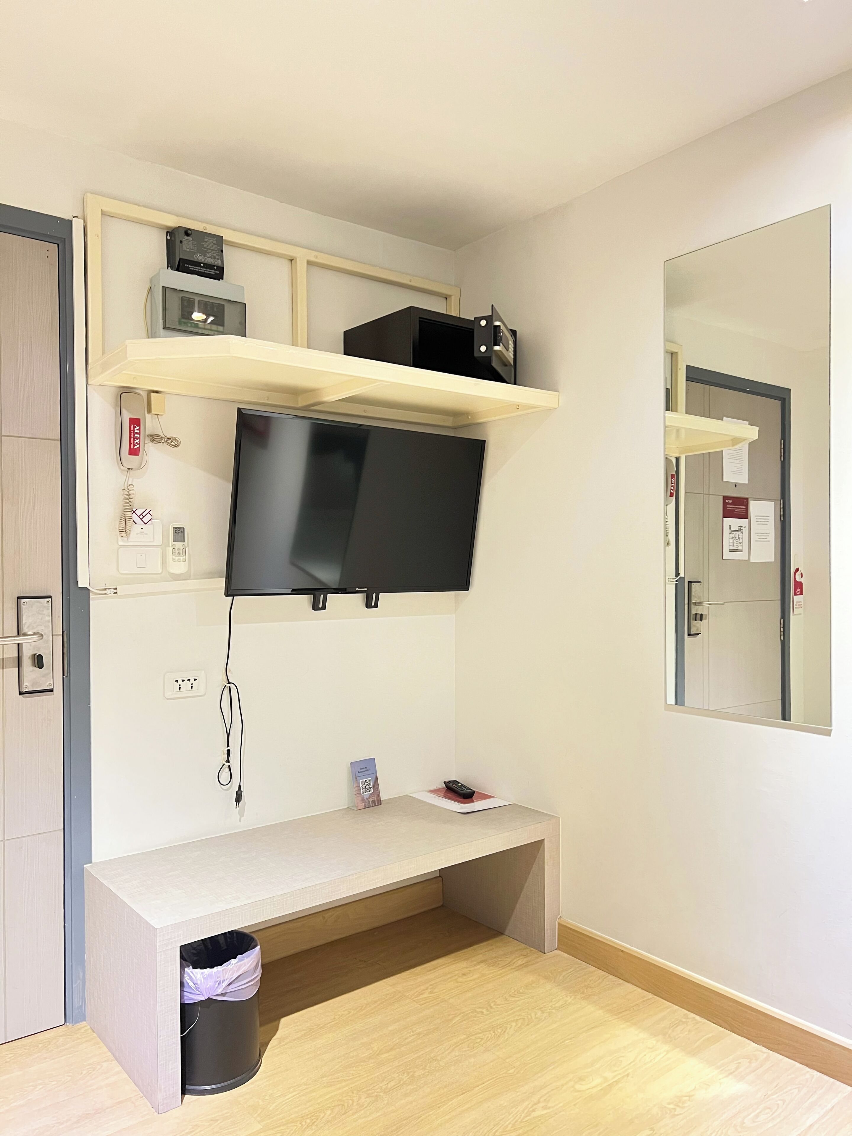 A room with a mounted TV, a desk, a trash bin, and a mirror on the wall.