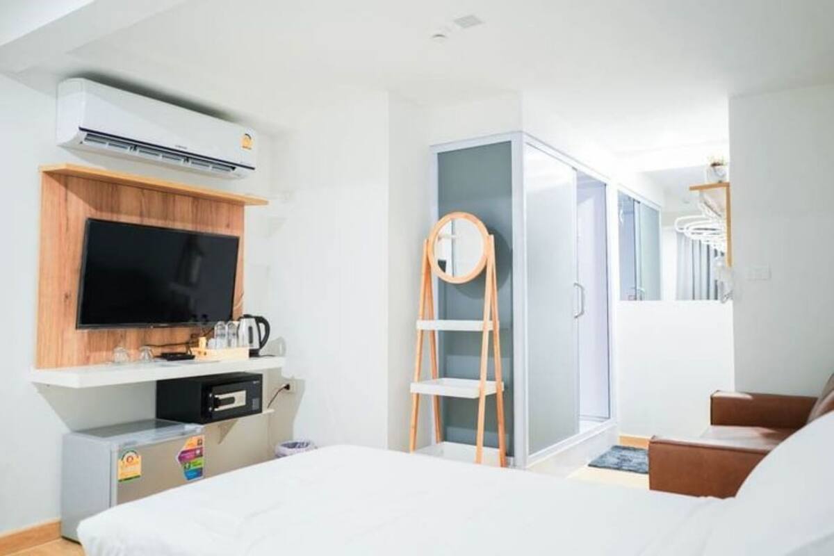 A modern hotel room with a bed, a TV, a mini-fridge, a microwave, a wooden shelf, a wardrobe, and a small seating area.