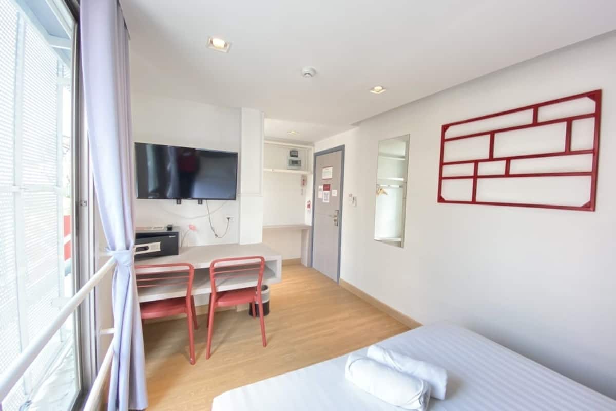 A hotel room with a bed, a desk with a chair, a TV mounted on the wall, and a window with curtains.
