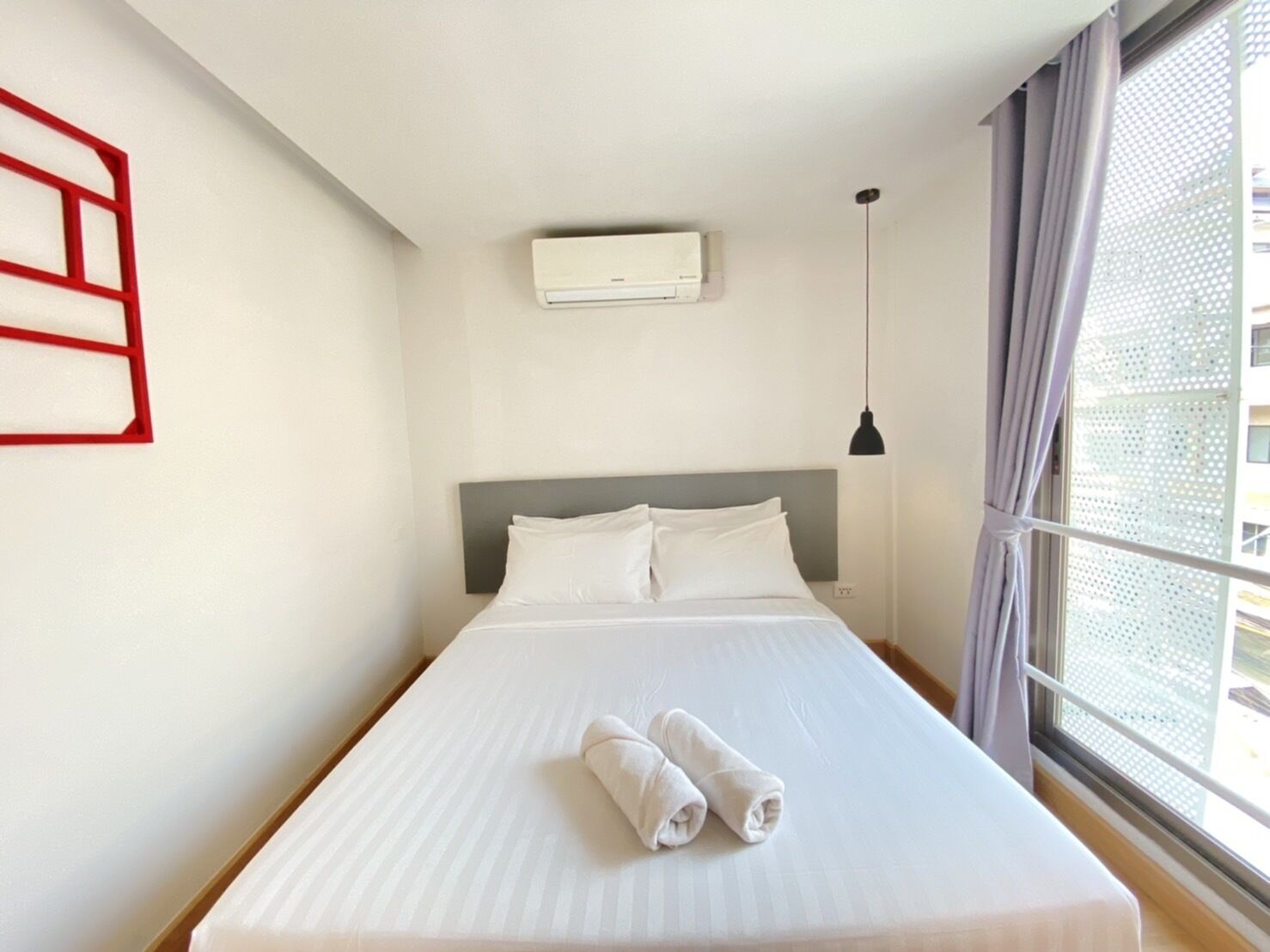 A hotel room with a bed, a wall-mounted air conditioner, a pendant light, and a window with curtains.