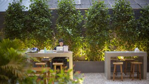 A patio area with a table and chairs, surrounded by lush greenery and a metal roof structure. - ALEXA Nimman - Hostel (Chiang Mai)