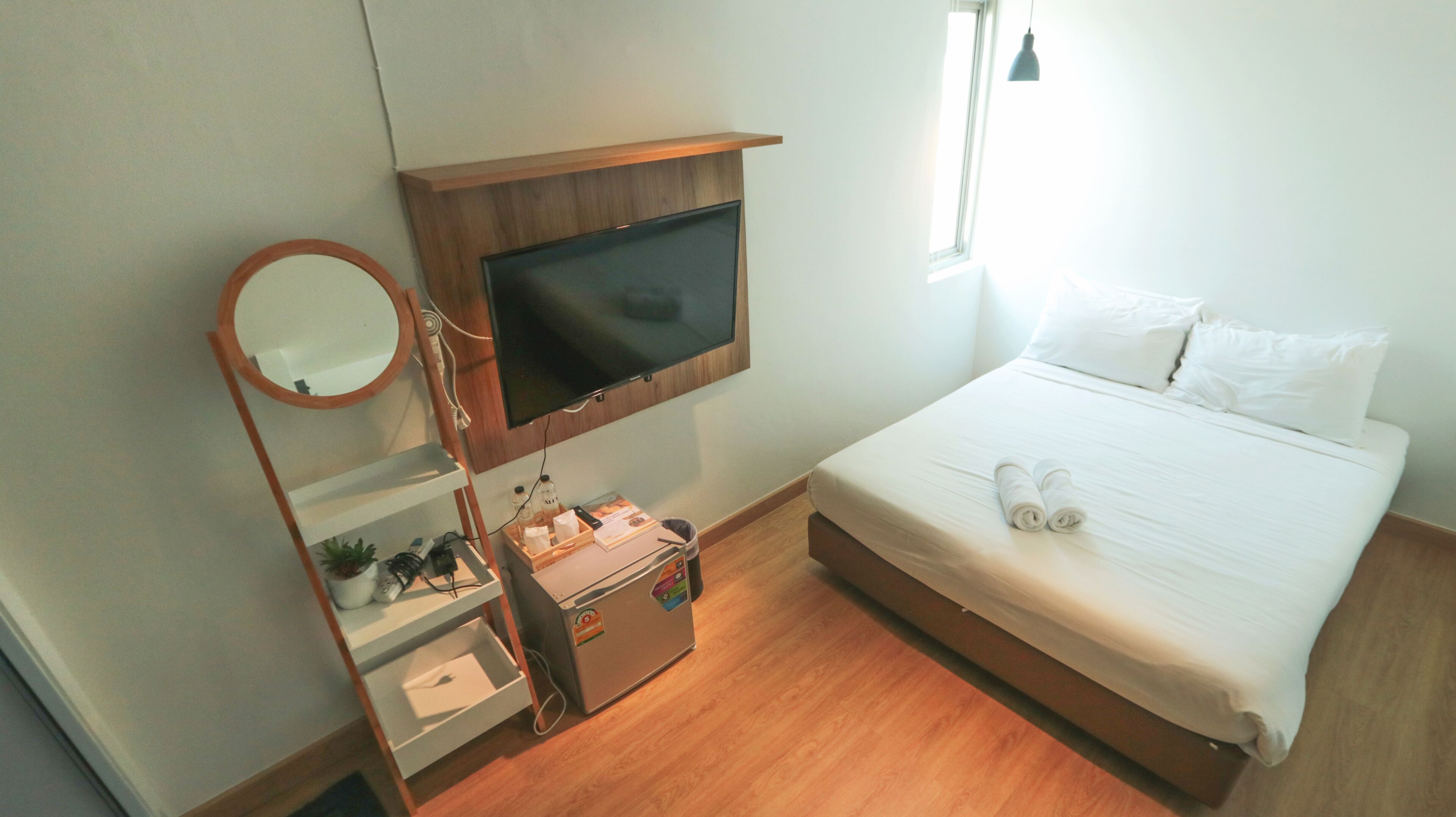 Ein Hotelzimmer mit Bett, Wandfernseher, Nachttisch mit Lampe und Fenster mit Tageslicht.