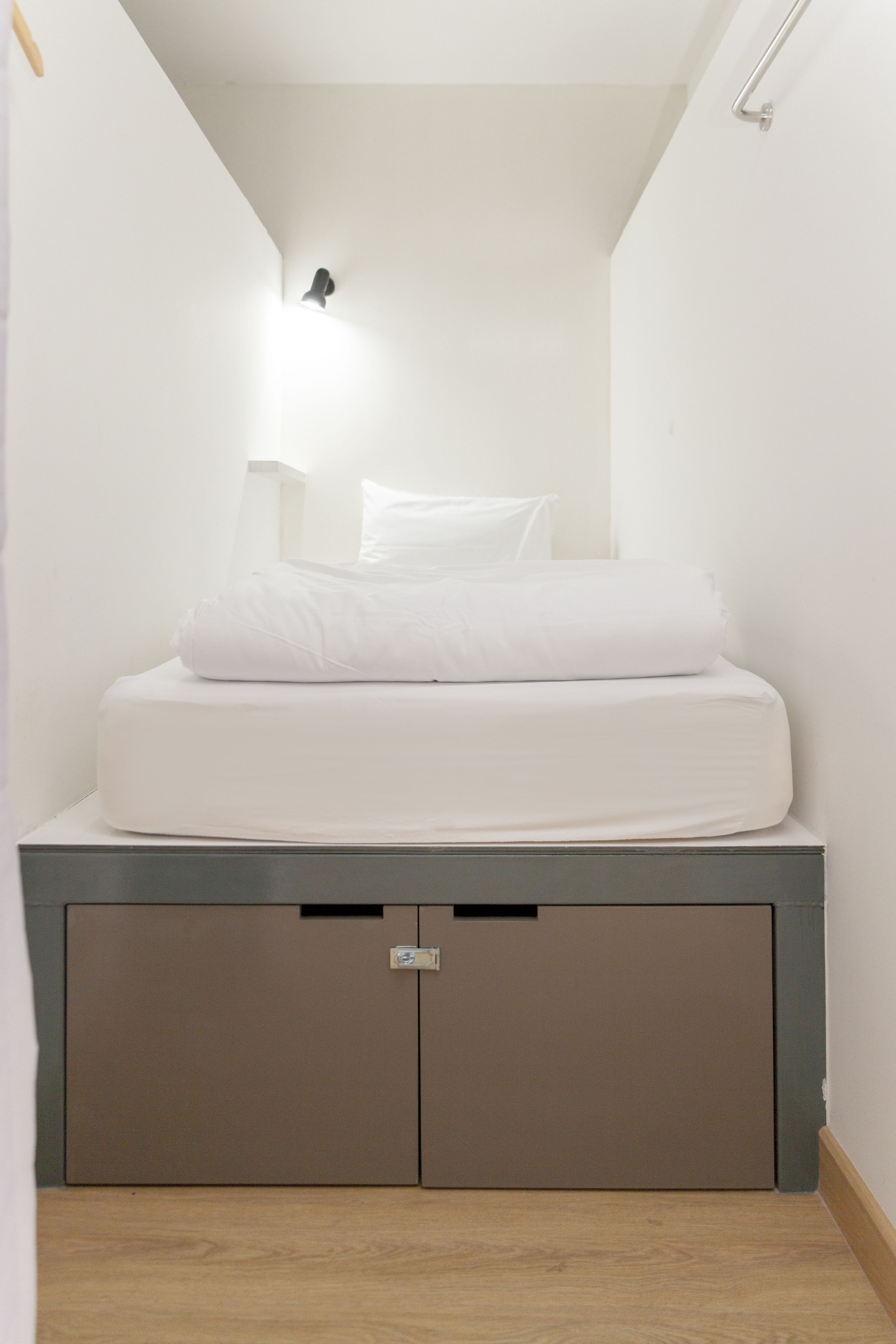 A small, narrow room with a bed, a nightstand, and a wall-mounted light fixture.