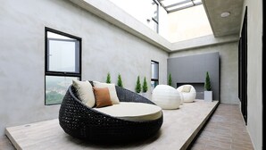 A modern interior with a round black bean bag chair, a white pouf, and a grey sofa. There are potted plants and a large window providing a view.