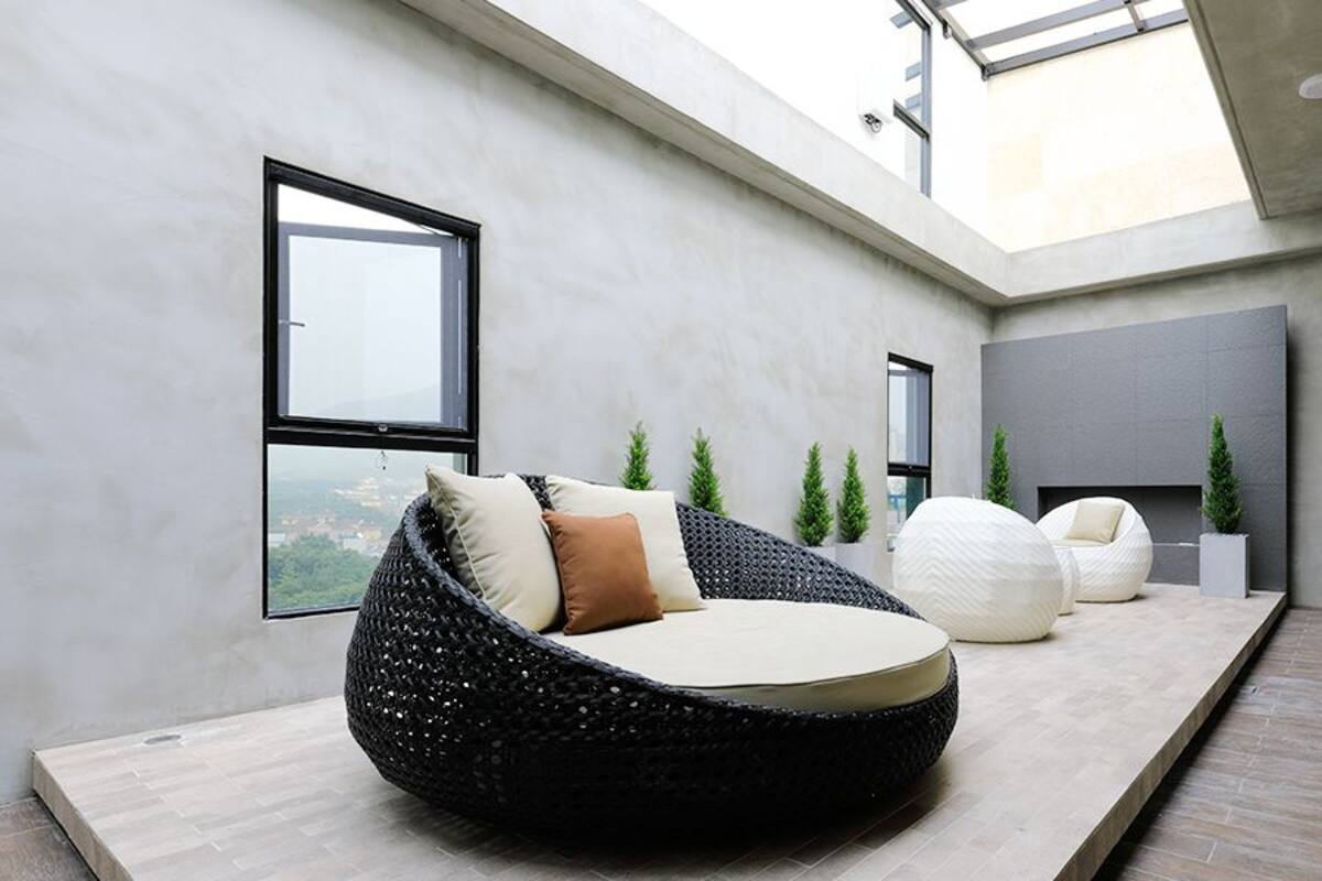 A modern interior with a round black bean bag chair, a white pouf, and a grey sofa. There are potted plants and a large window providing a view.