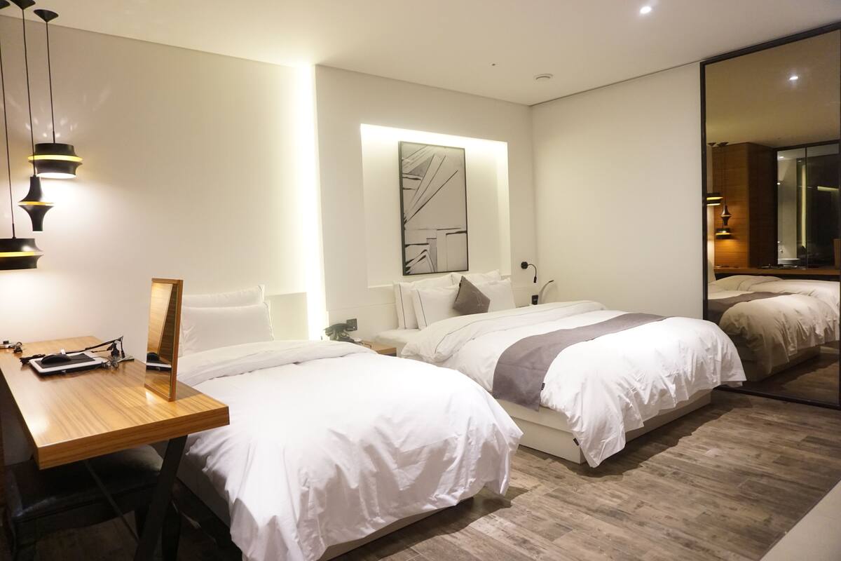 A modern hotel room with two beds, a wooden desk, a large mirror, and a contemporary art piece on the wall.