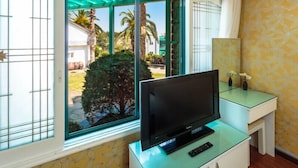 Une pièce avec un téléviseur posé sur une table en verre, une fenêtre donnant sur un paysage verdoyant et un bâtiment à l’extérieur, et un mur orné d’un motif.