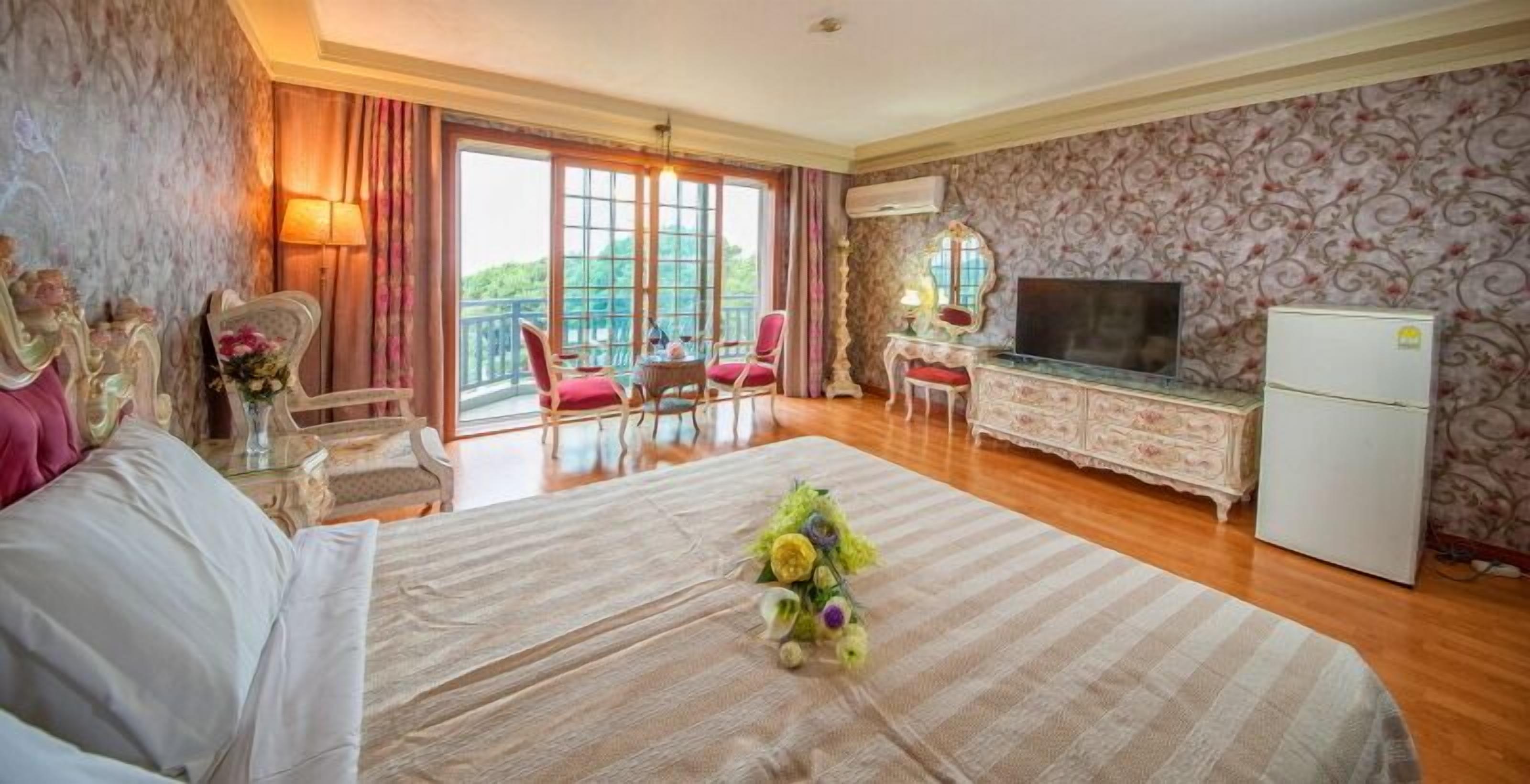 A hotel room with a large bed, a television, a refrigerator, a balcony with red chairs, and a view of greenery.