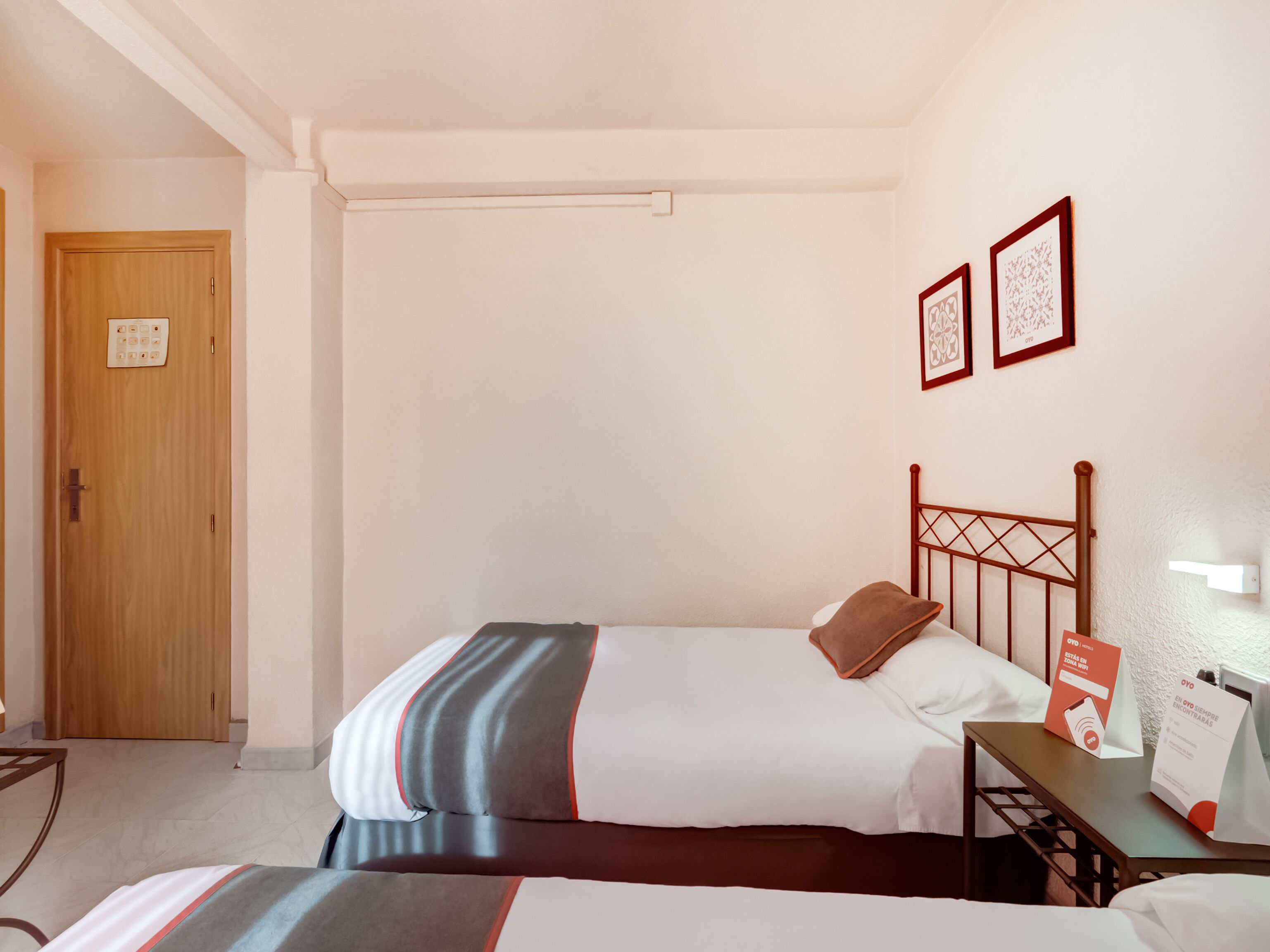 Een hotelkamer met twee bedden, een houten deur, een klein tafeltje met een bordje en twee ingelijste foto's aan de muur.
