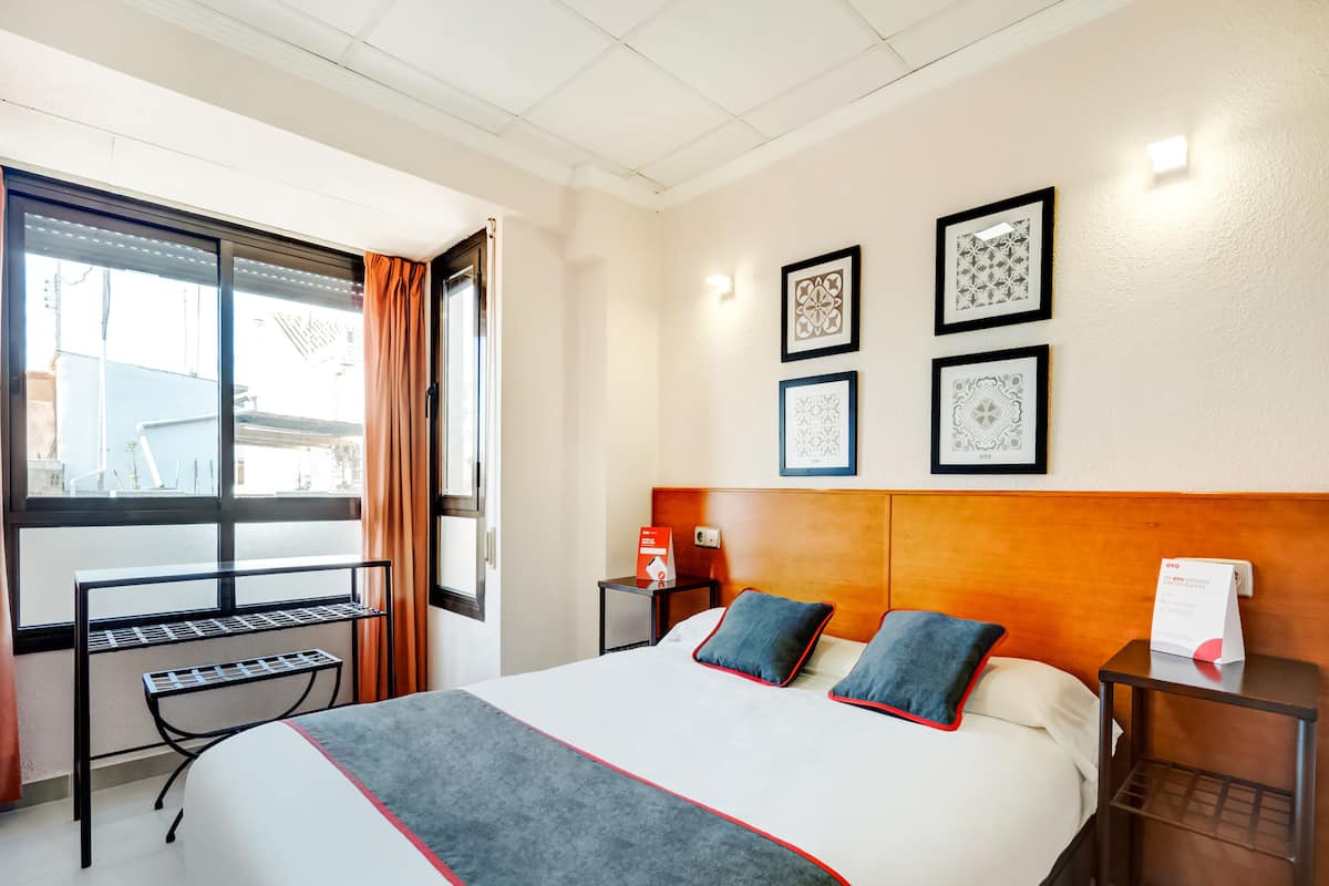 A hotel room with a large bed, a window with curtains, a bedside table with a lamp, and framed artwork on the wall.