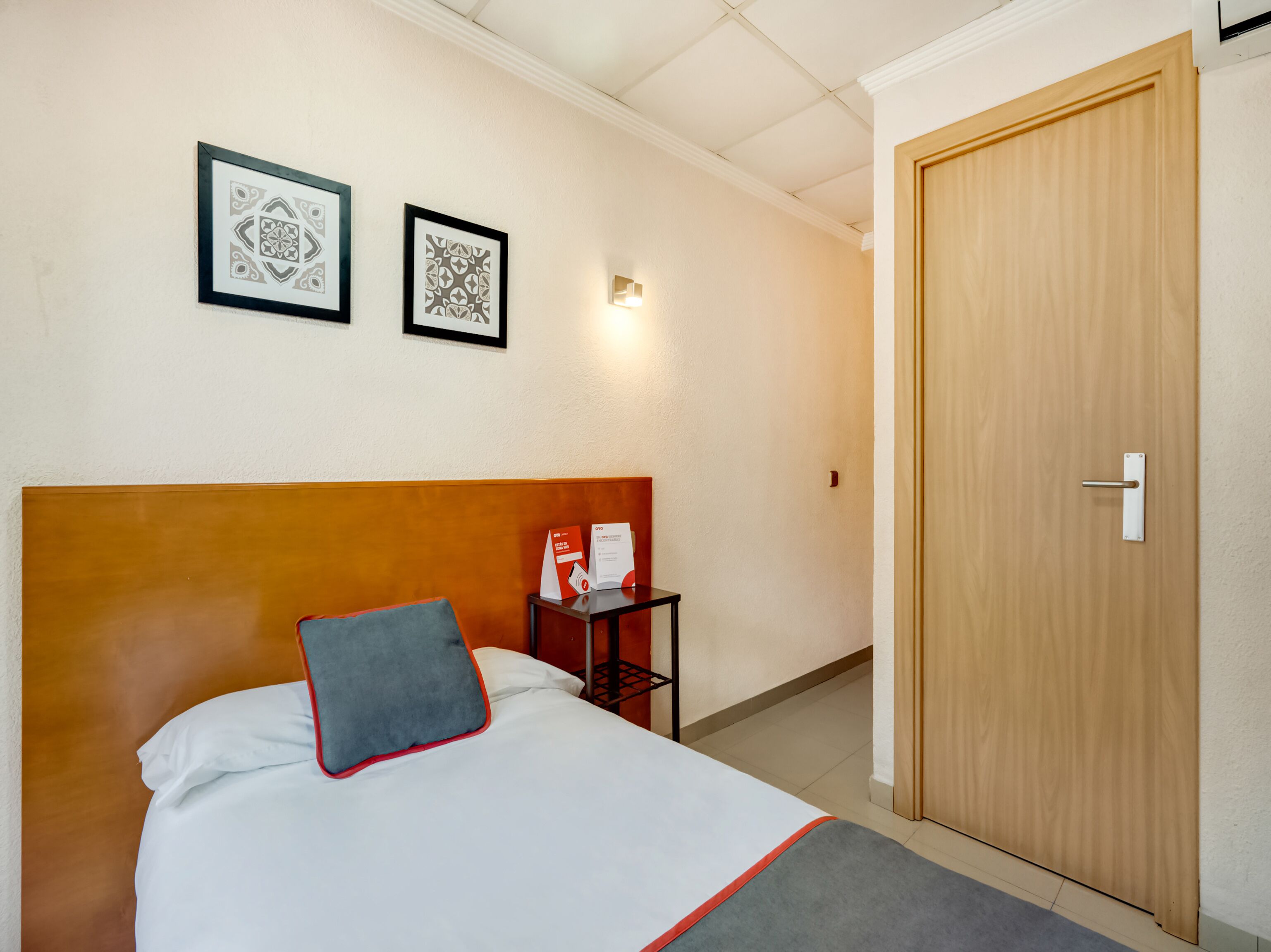 Ein Hotelzimmer mit einem hölzernen Kopfteil, einem Bett mit weißer Bettwäsche und einem grauen Kopfkissen, zwei gerahmten Bildern an der Wand, einem kleinen Tisch mit einem rot-weißen Schild und einer Holztür.