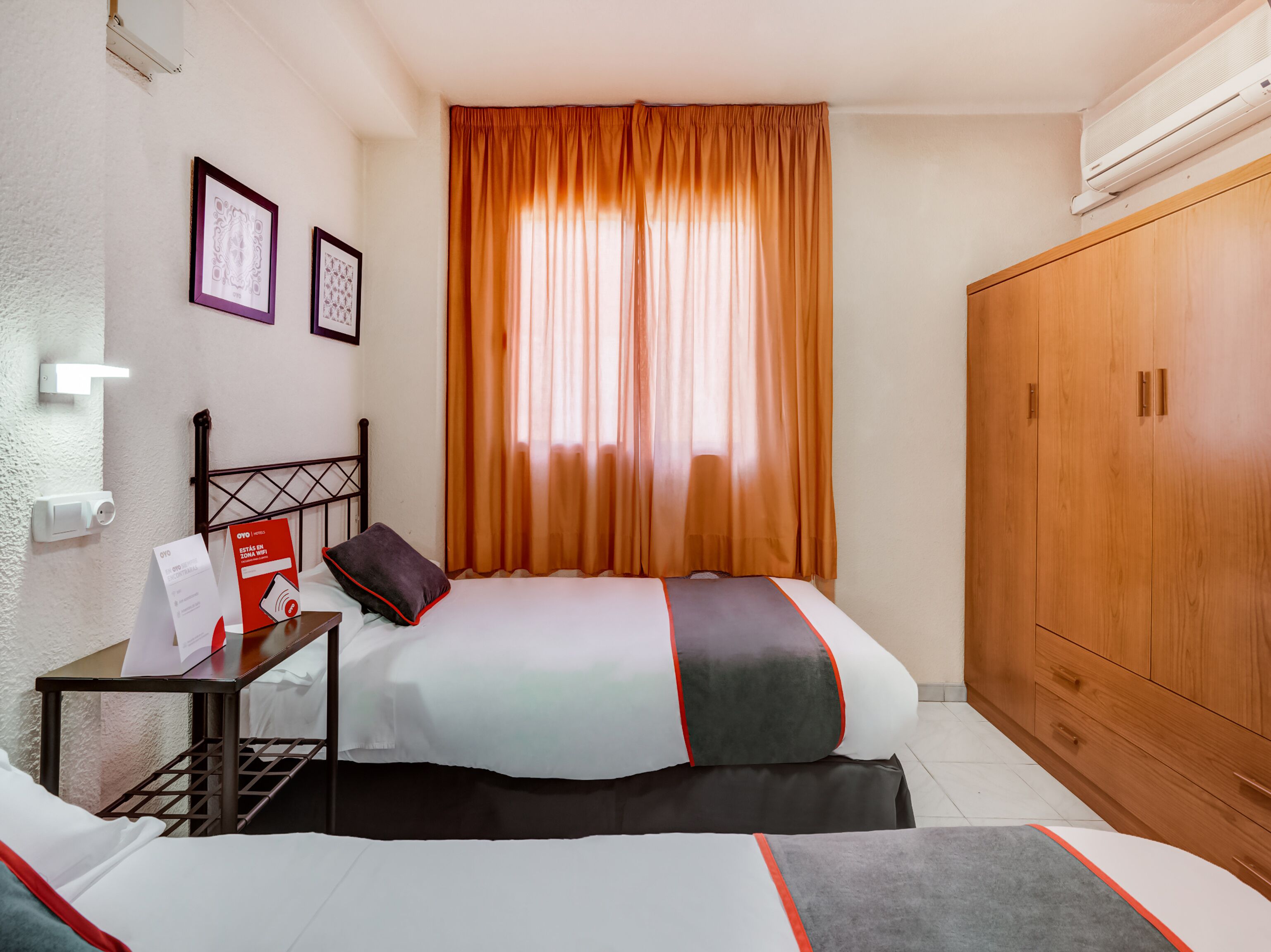 Habitació d'hotel amb dos llits, un armari de fusta, una tauleta de nit amb una làmpada i una finestra amb cortines taronges.