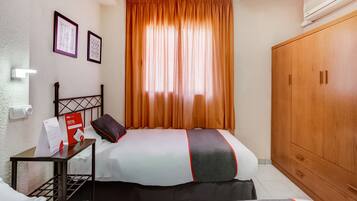 Habitació d'hotel amb dos llits, un armari de fusta, una tauleta de nit amb una làmpada i una finestra amb cortines taronges.