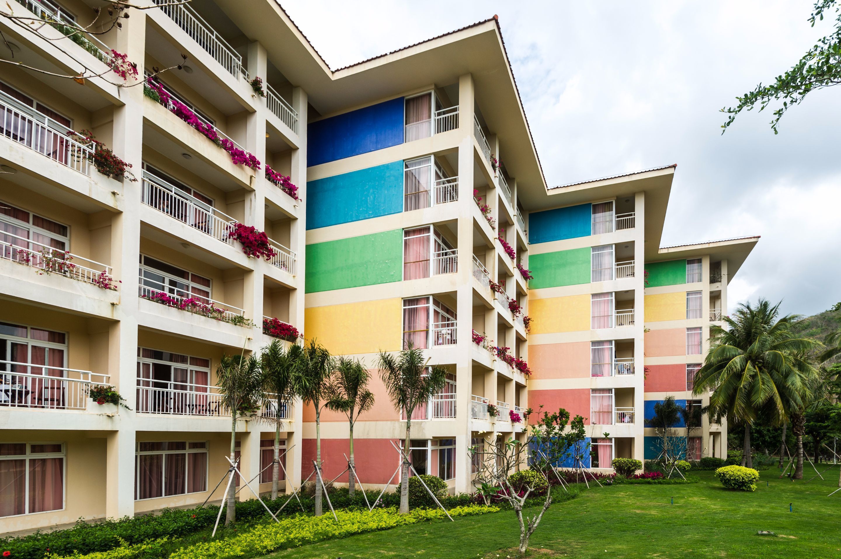 A multi-story hotel with balconies adorned with flowers, a green lawn, and palm trees.