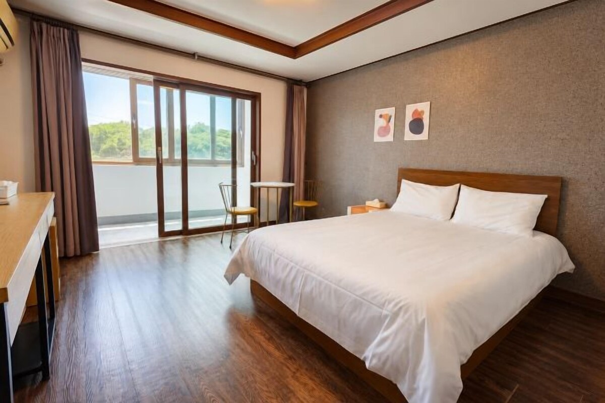 A hotel room with a large bed, a desk, a chair, and a balcony with curtains.