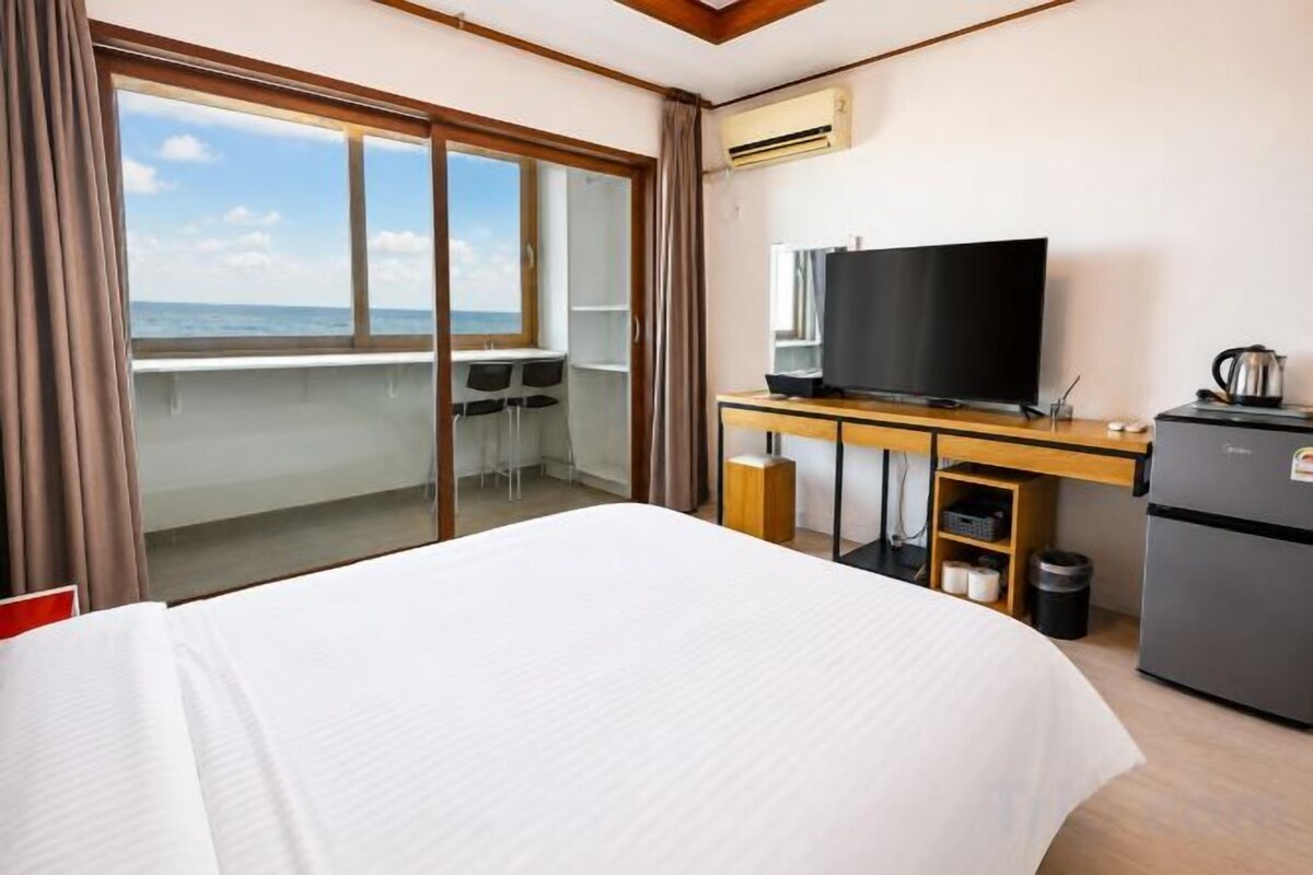 A hotel room with a bed, a desk with a television, a refrigerator, and a view of the sea.