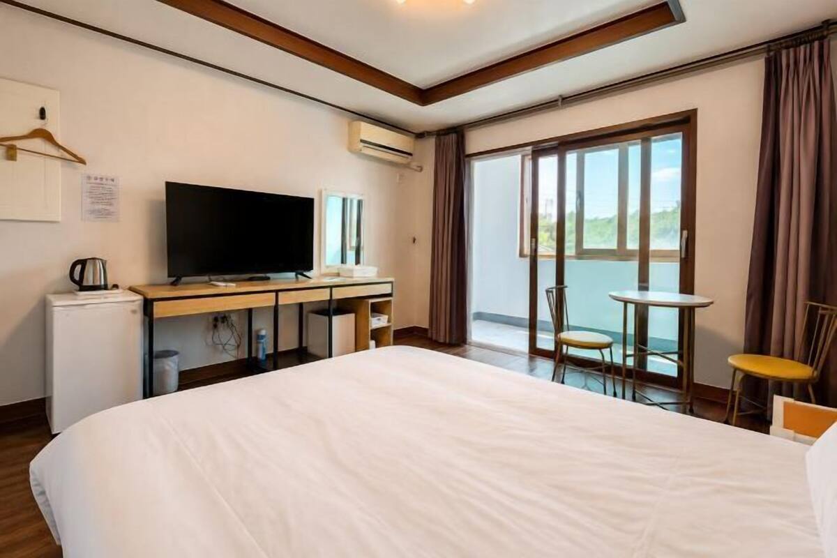 A hotel room with a large bed, a desk with a television, a small refrigerator, and a balcony with chairs.