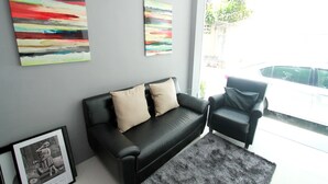 A modern living room with a black leather sofa, two beige cushions, and a black armchair. There are two abstract paintings on the wall and a framed photograph on the floor. - Coco at 20 (Bangkok)