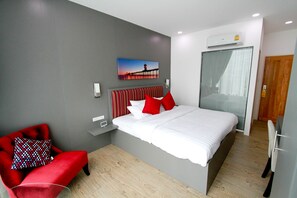 A modern hotel room with a large bed, a red armchair, a painting on the wall, and a window with blinds. - Coco at 20 (Bangkok)