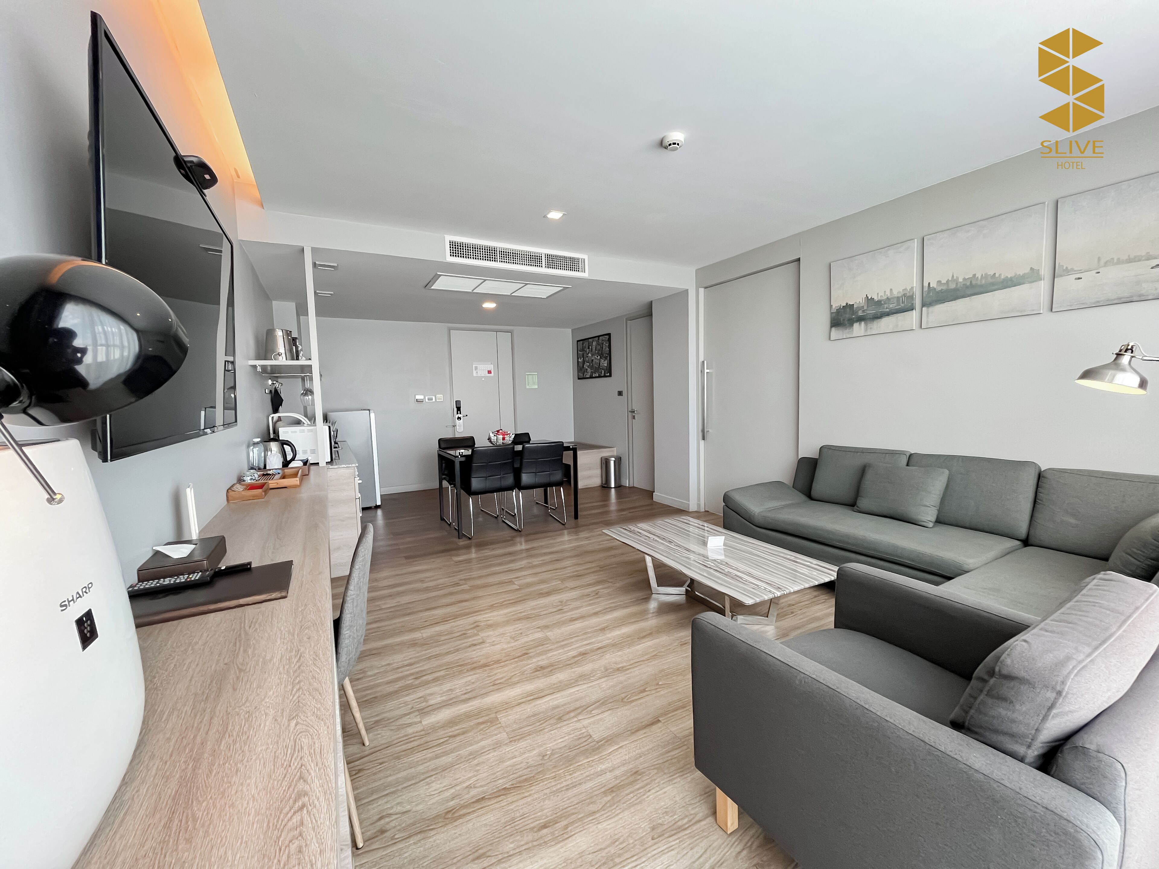 A modern hotel room with a grey sofa, a dining area, and a flat-screen TV.