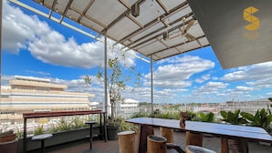 A rooftop terrace with a wooden table and stools, potted plants, and a view of the cityscape.