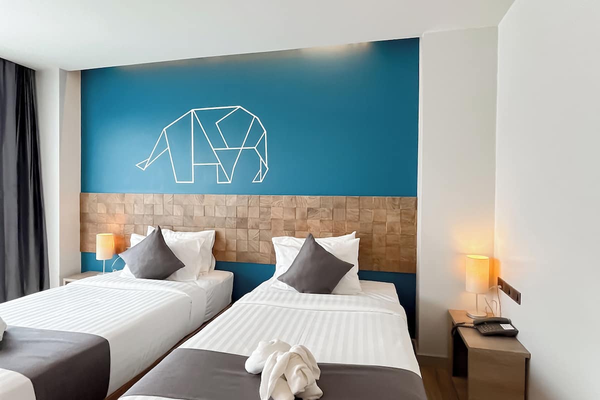 Ein Hotelzimmer mit zwei Betten, einer blauen Wand mit geometrischem Muster, einem Holzkopfteil, Nachttischlampen und einem Bademantel auf dem Bett.