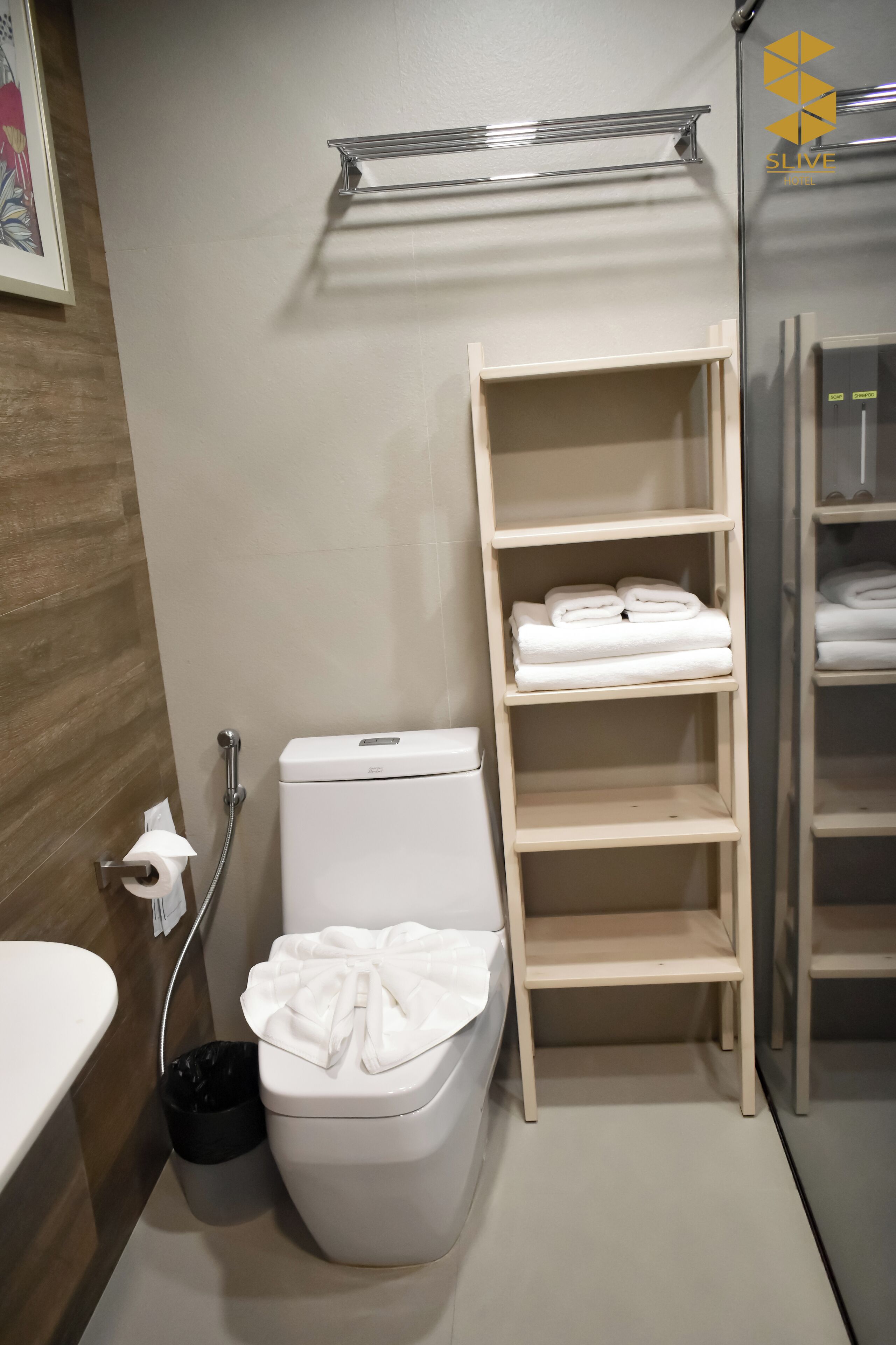 Een badkamer met toilet, een plank met opgevouwen handdoeken, een prullenbak en een douchehoek.