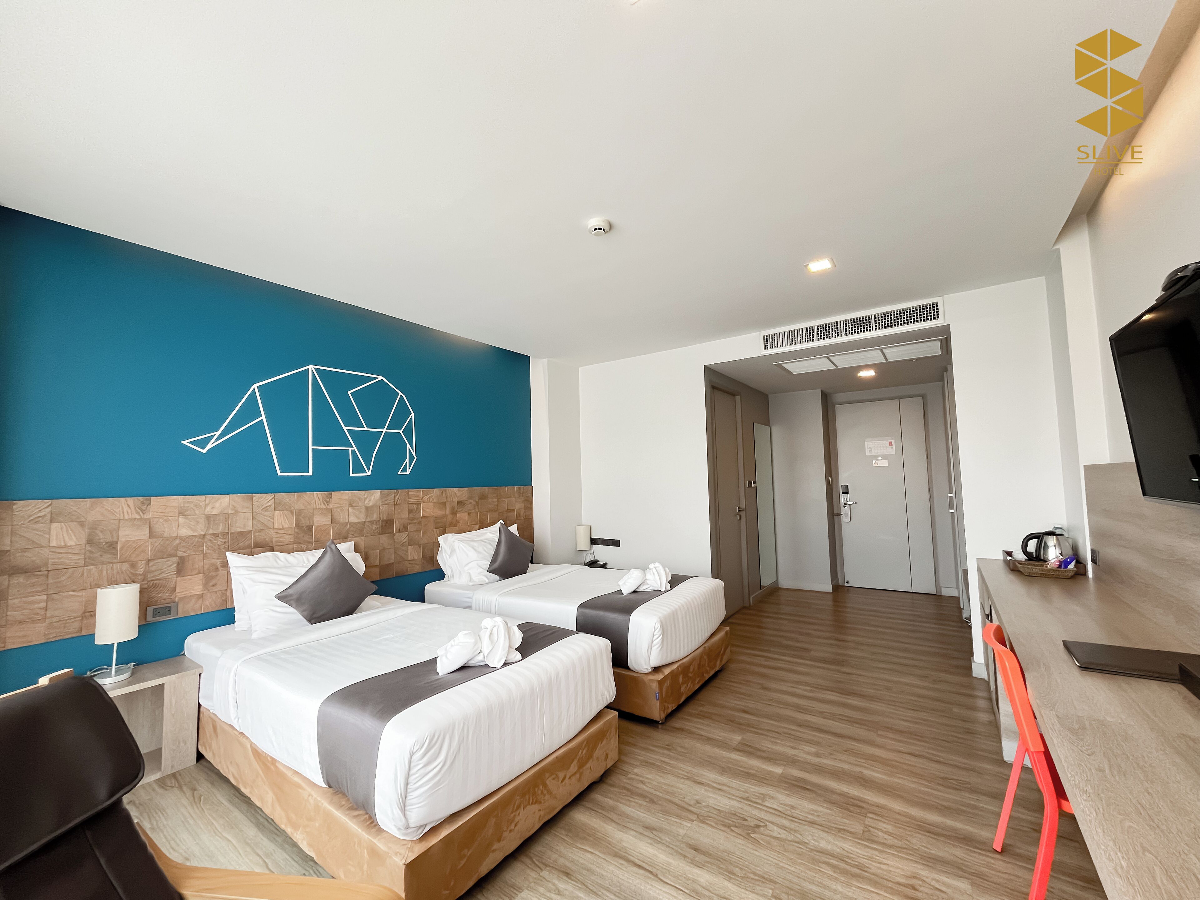 Habitación de hotel con dos camas, cabecero de madera, pared azul de acento, piso de madera, escritorio con silla y televisor.