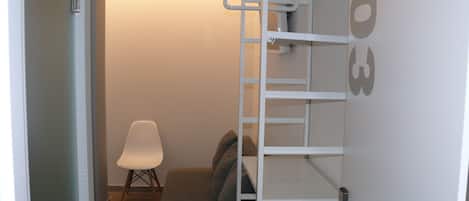 Habitación pequeña con un sofá gris, una silla blanca y una estantería blanca con escalera.