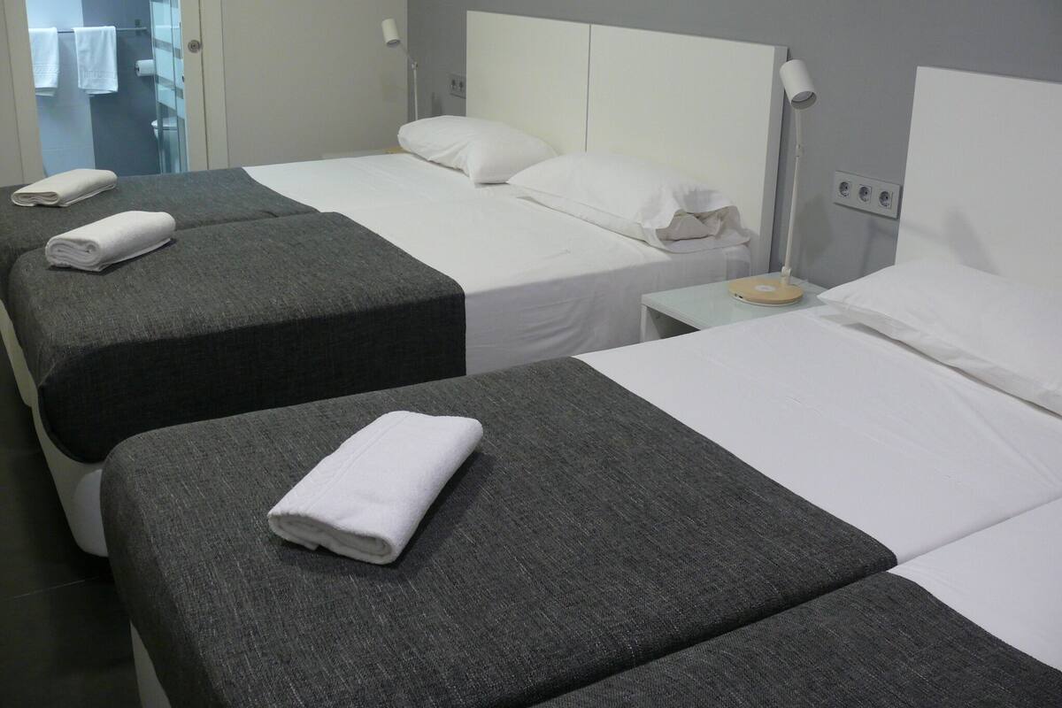 A hotel room with two beds, each with a grey headboard and white bedding. There are towels on the beds, a bedside table with a lamp, and a small table with a scale.