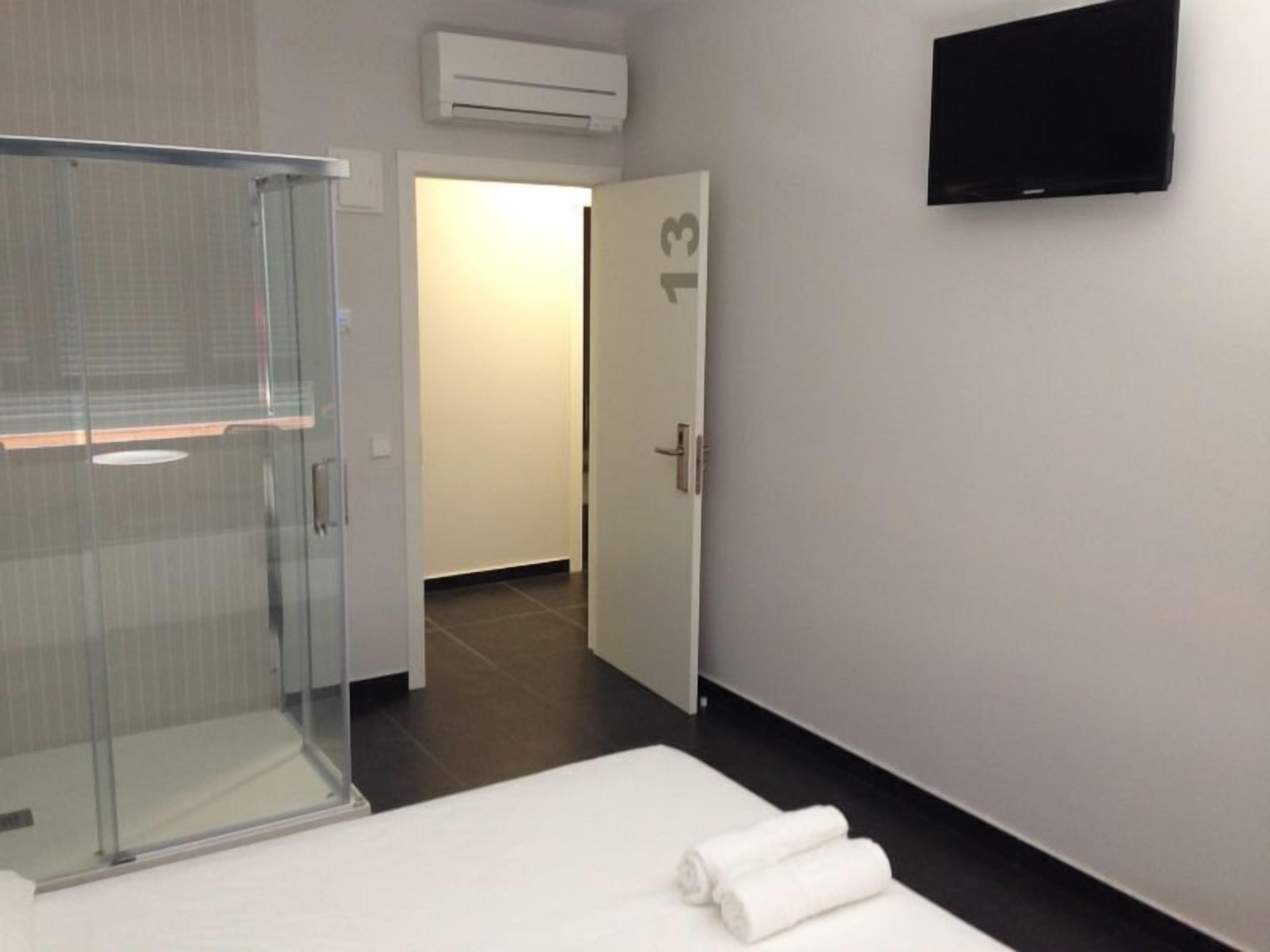 A hotel room with a glass shower enclosure, a bed with white linens, a flat-screen TV mounted on the wall, and a door with the number 13.
