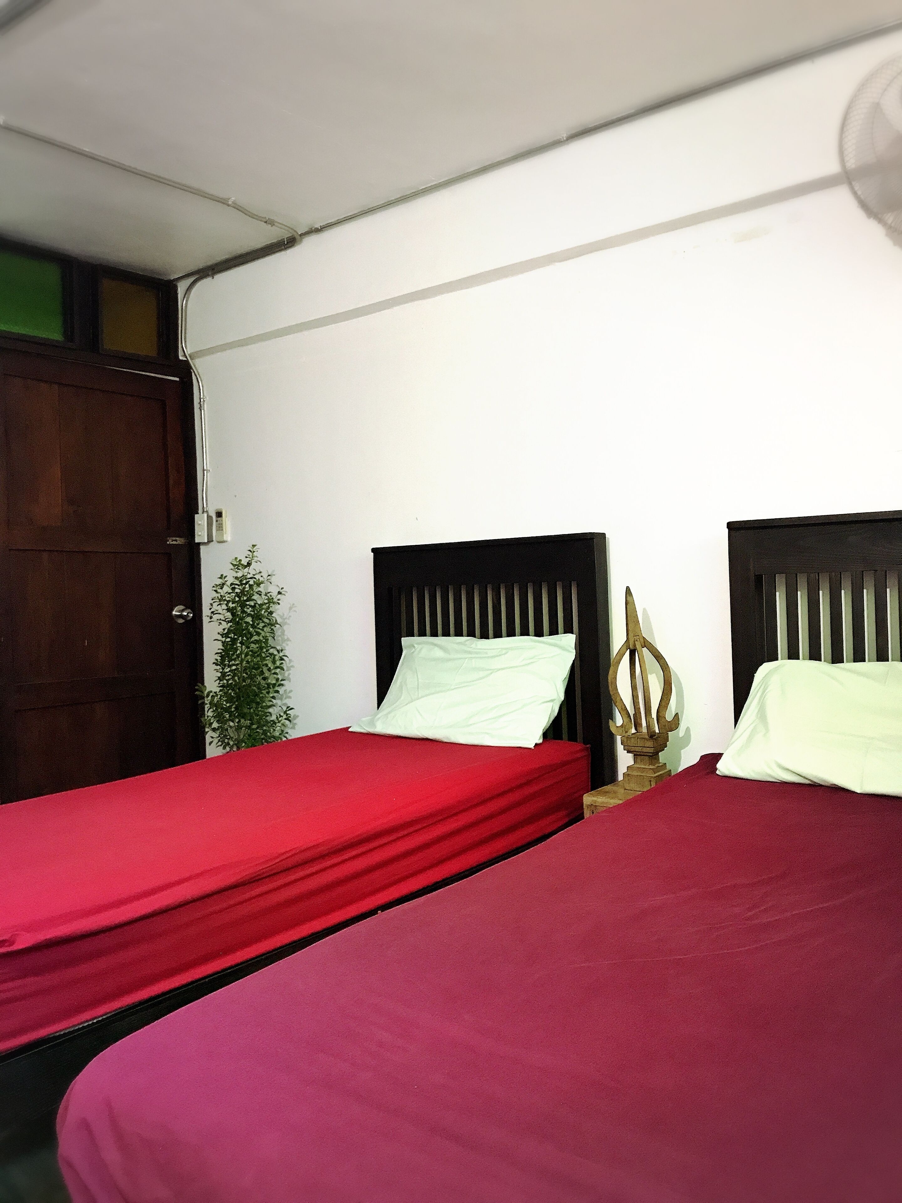 A room with two beds, each with a wooden headboard and a red bedspread. There is a wooden door and a small potted plant in the corner.