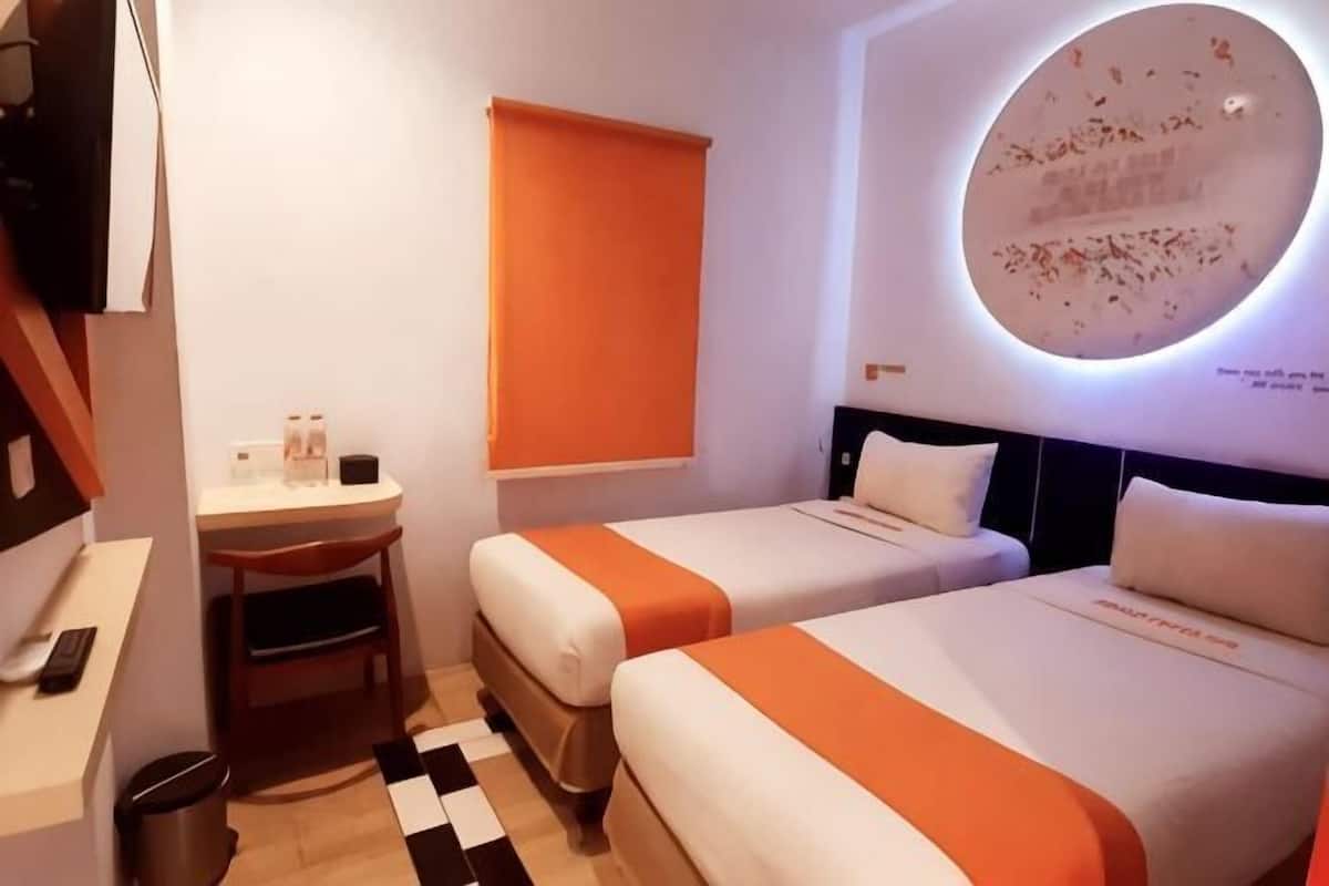 A hotel room with two beds, a desk, a trash bin, and a wall-mounted TV.