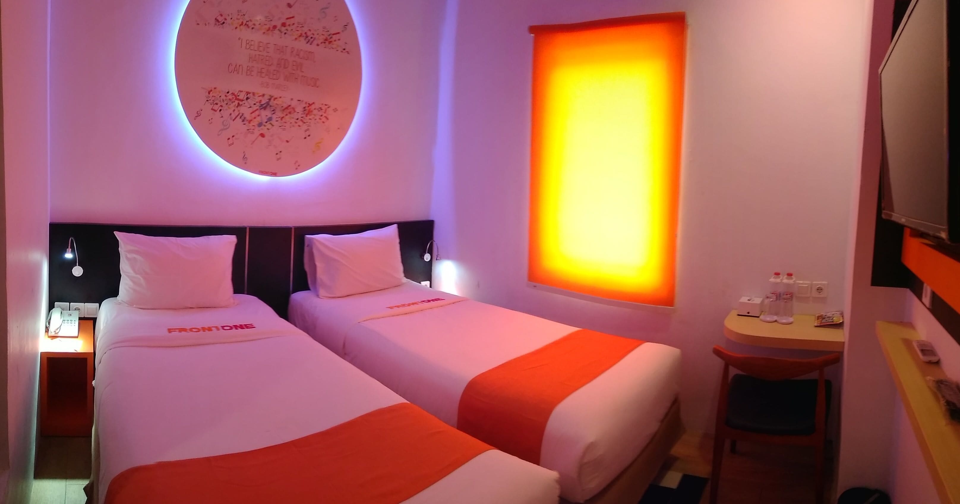 Une chambre d’hôtel avec deux lits, un élément de décoration circulaire au mur, une fenêtre laissant entrer une lumière chaleureuse et une petite table avec une chaise.