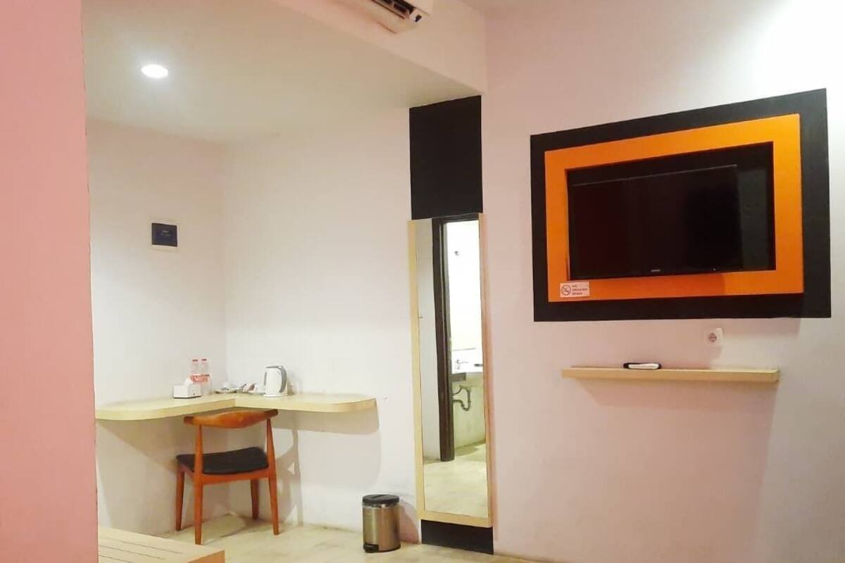 A hotel room with a bed, a desk with a chair, a small trash bin, a mounted TV, and an air conditioning unit.