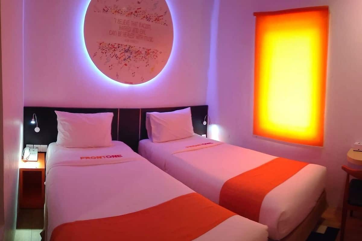 A hotel room with two beds, a wall-mounted TV, a small table with a chair, and a circular light fixture with text.