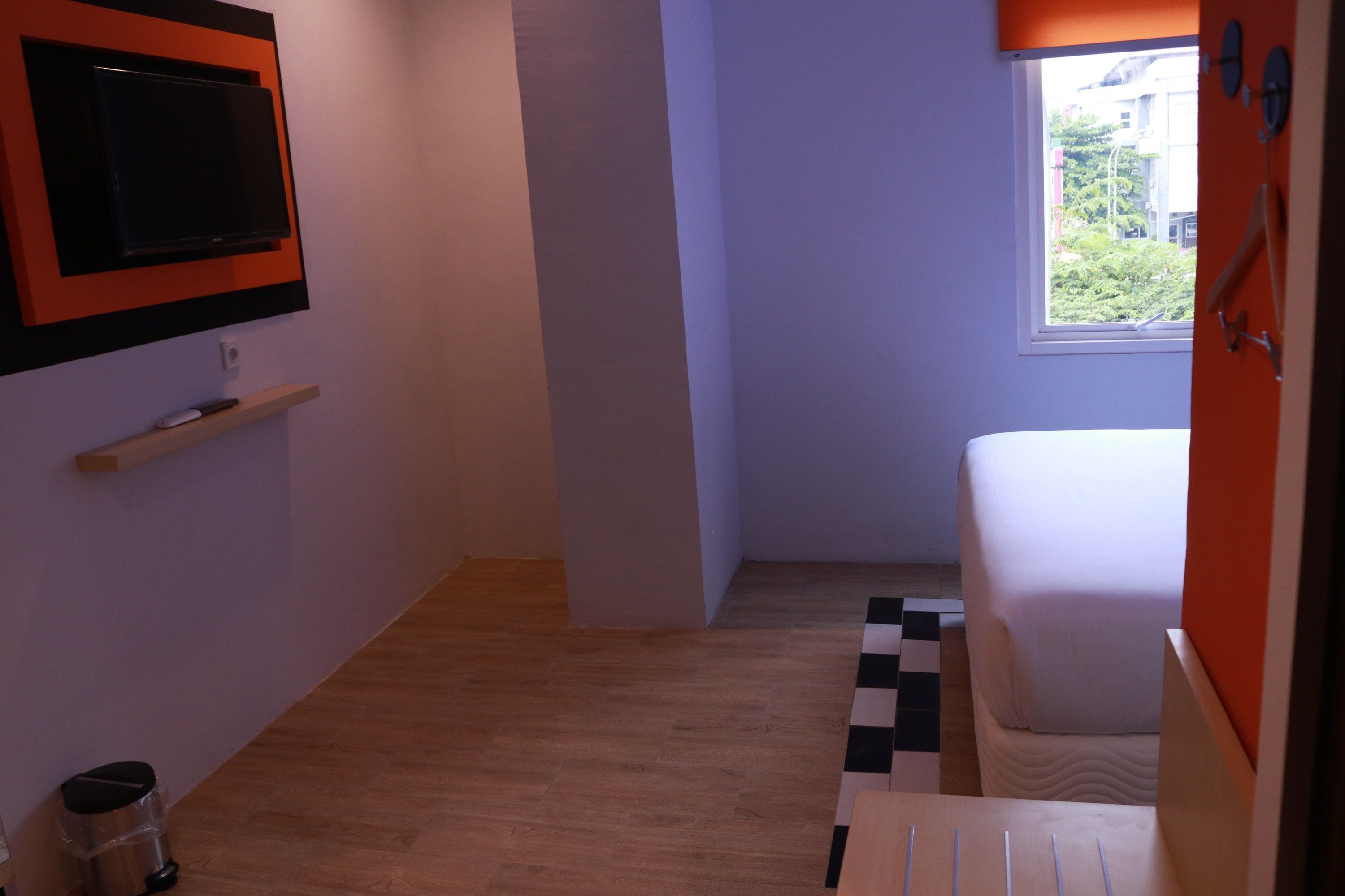 Una habitación pequeña y moderna con una cama, un televisor montado en la pared, una ventana con vistas a zonas verdes y un cubo de basura.