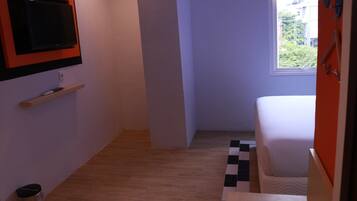 Una habitación pequeña y moderna con una cama, un televisor montado en la pared, una ventana con vistas a zonas verdes y un cubo de basura.