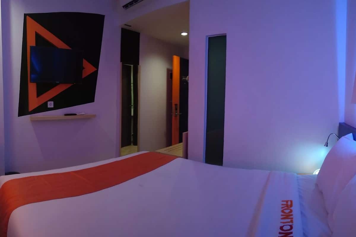 A hotel room with a bed, a desk with a chair, a wall-mounted TV, and a door leading to another room.