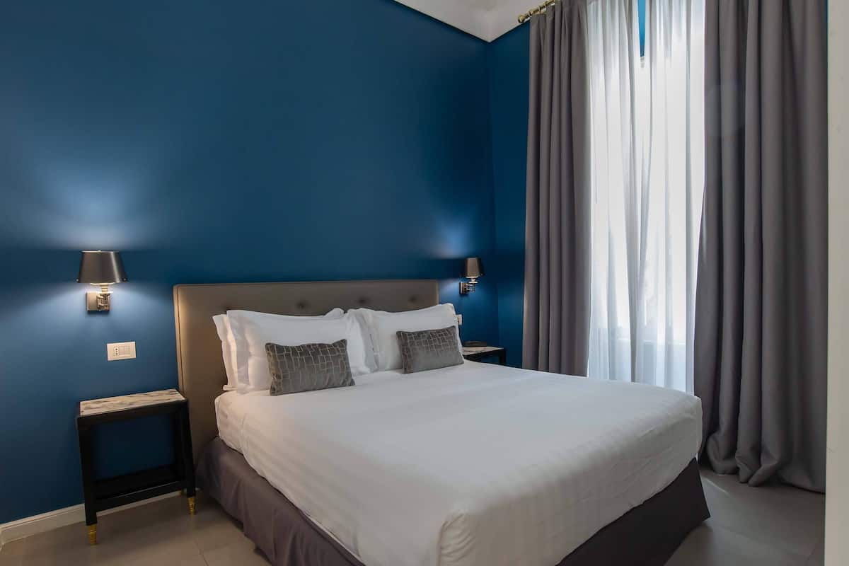 A neatly made bed with white linens and gray pillows, a bedside table with a lamp, and a window with curtains.