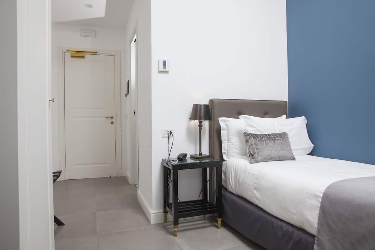 A hotel room with a bed, bedside table, lamp, and a door leading to another room.