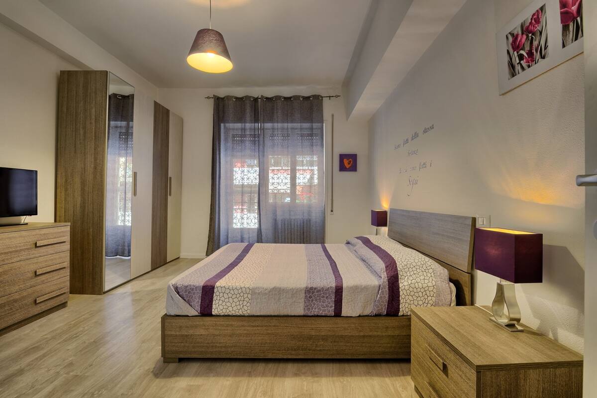 A modern bedroom with a wooden bed, a bedside table with a lamp, a TV on a wooden dresser, and a wardrobe with a mirror.