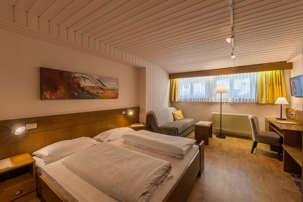A hotel room with two beds, a desk, a chair, a lamp, a radiator, and a painting on the wall. - Hotel Mondschein (Sesto)