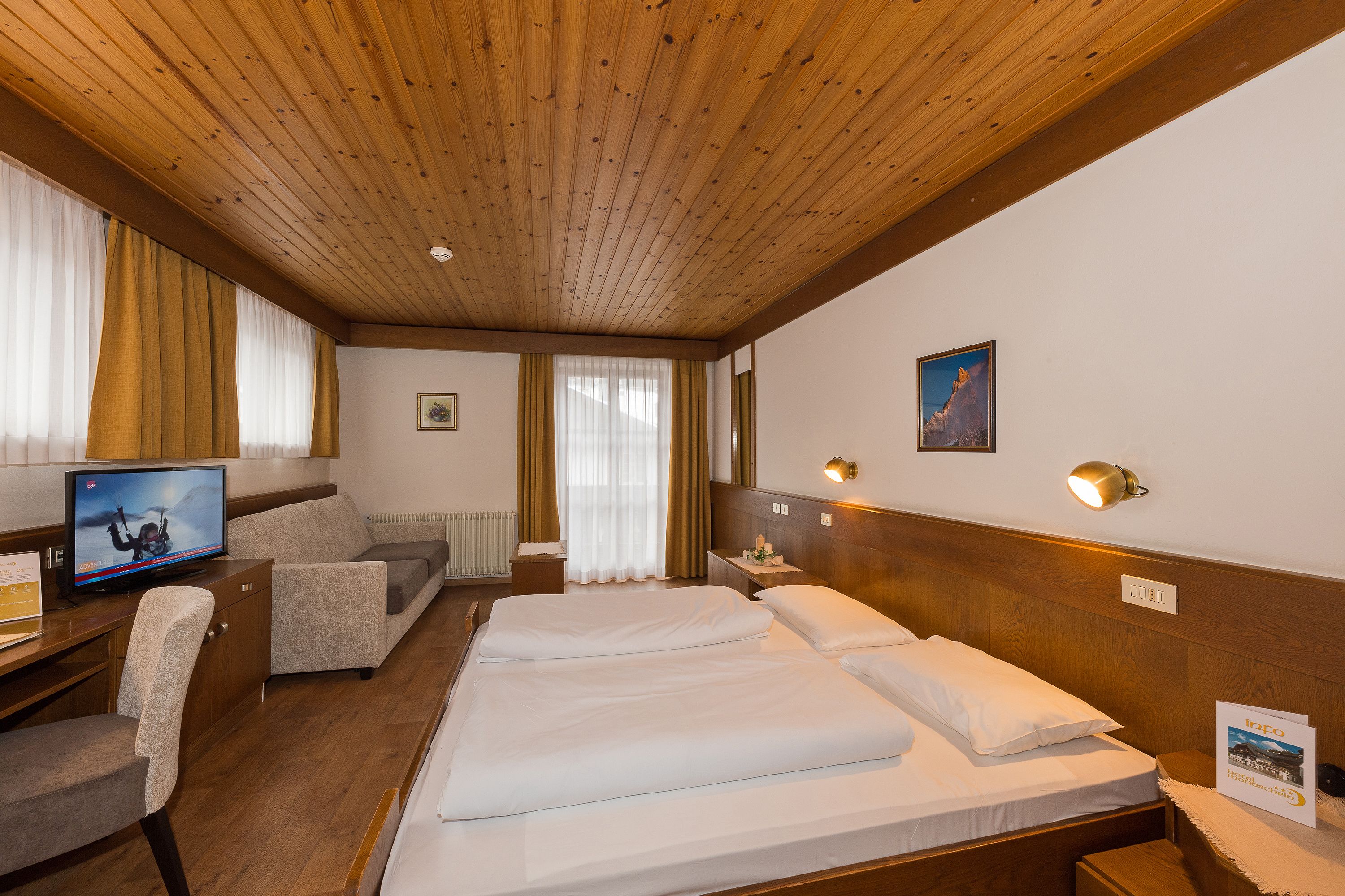 Ein Hotelzimmer mit Holzboden, einem Bett mit weißer Bettwäsche, einer Couch, einem Schreibtisch mit Fernseher und einem Nachttisch mit Lampe.