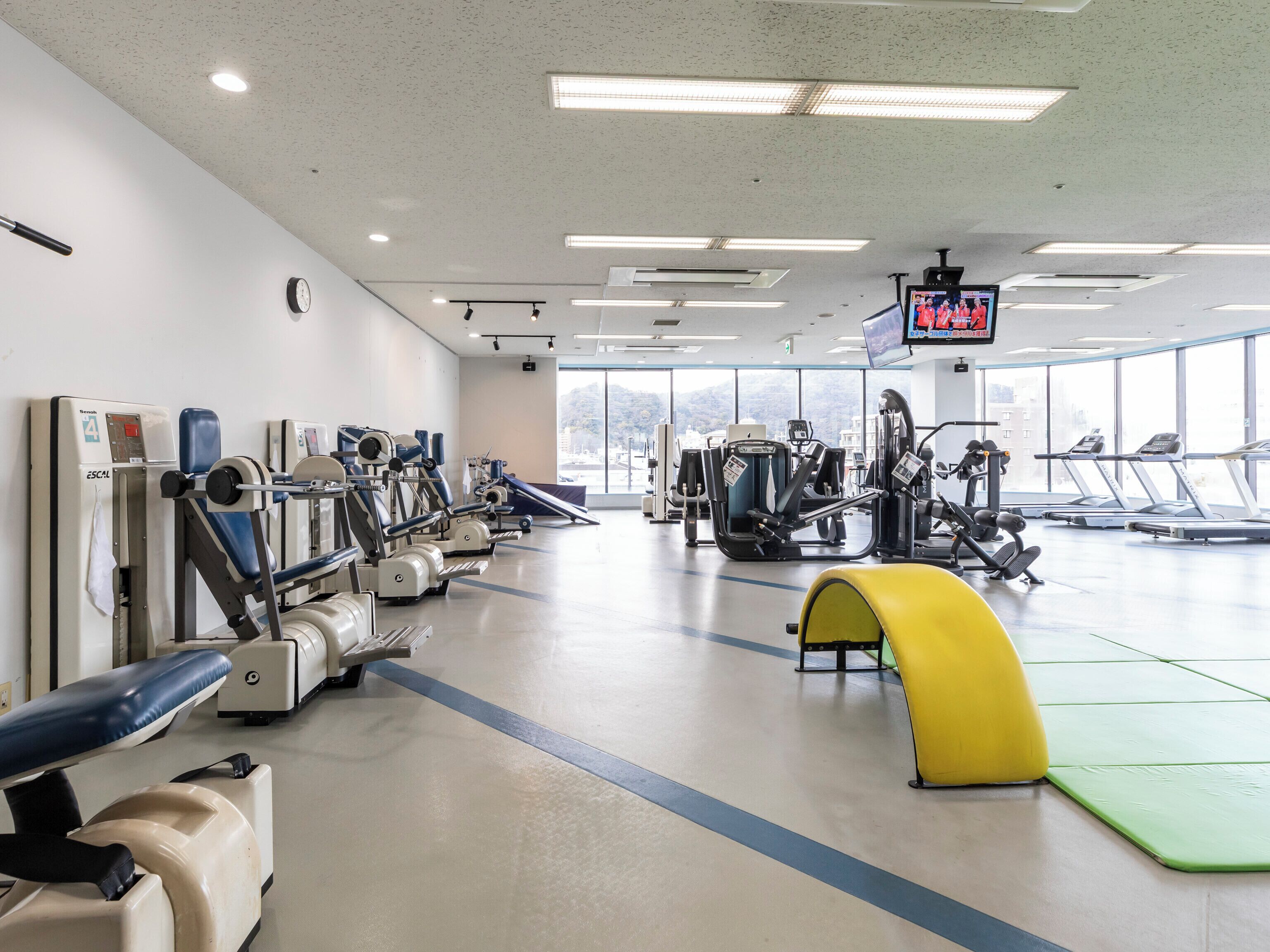 A gym with various exercise machines, treadmills, and a large window providing a view of the city.
