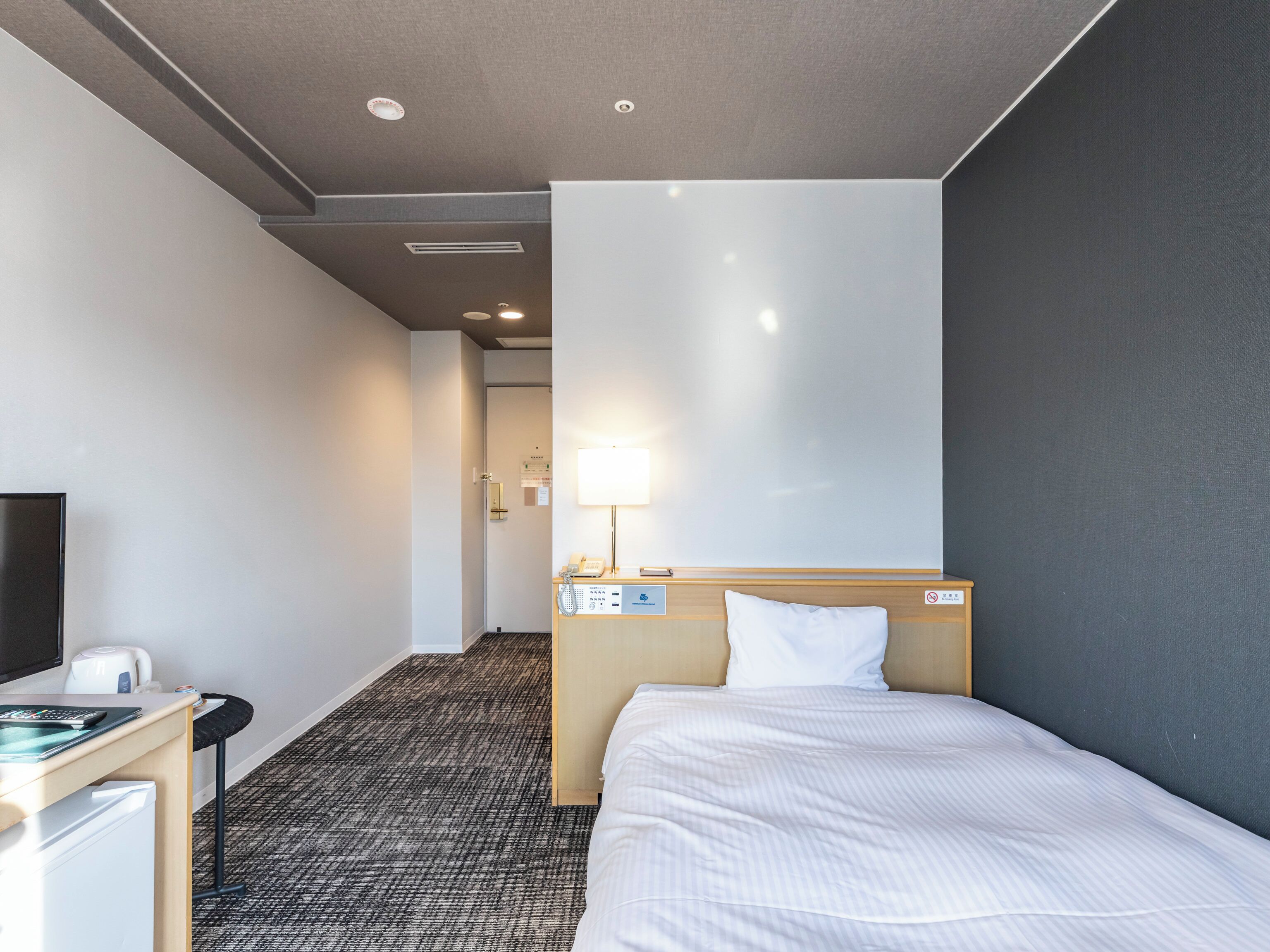 A hotel room with a bed, a desk with a computer and a coffee maker, a lamp, and a door leading to a hallway.