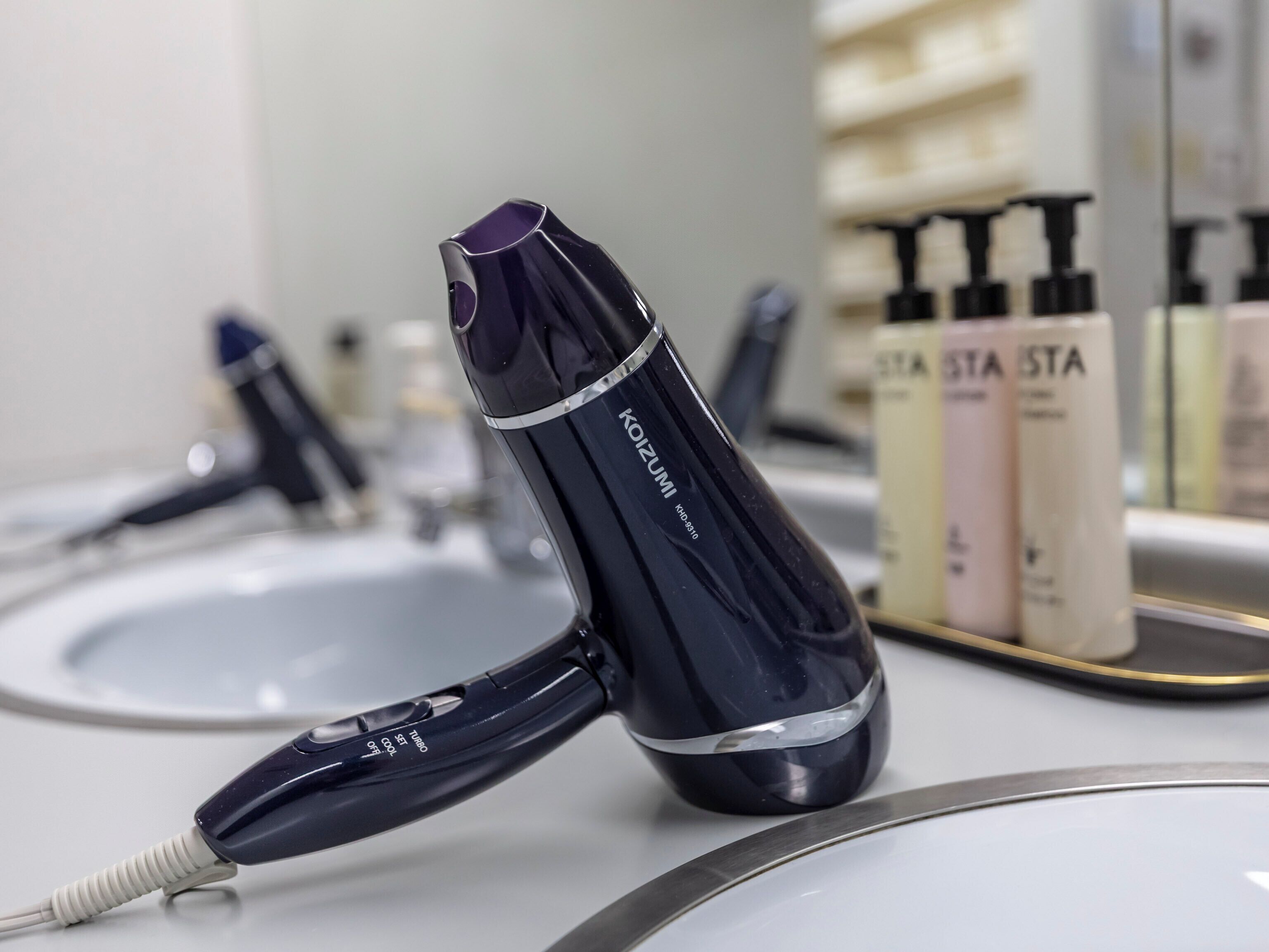 Un secador de pelo sobre una encimera, con un espejo que refleja un expositor de productos para el cabello.