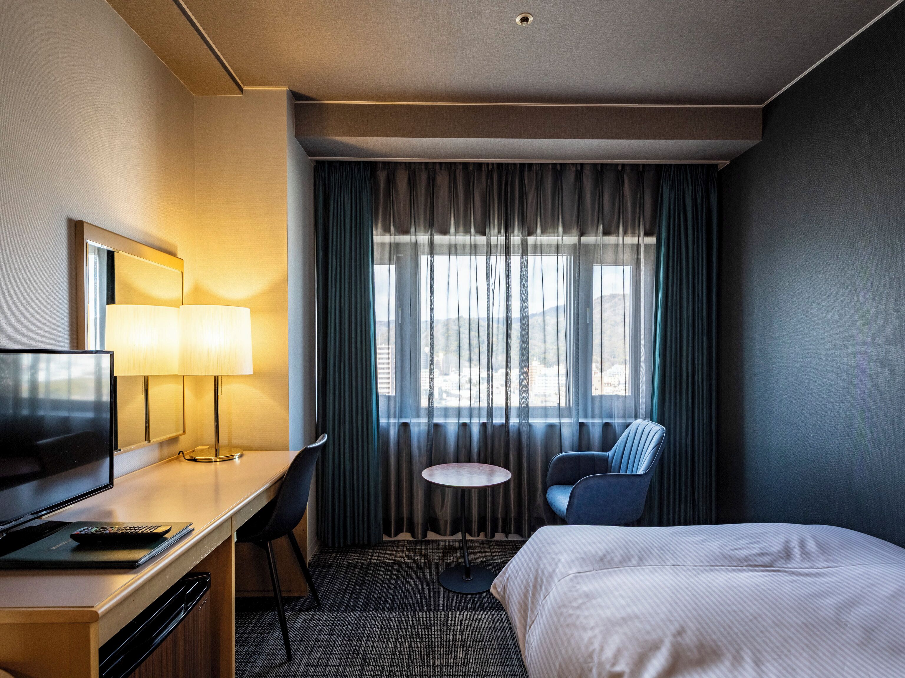 Une chambre d’hôtel avec un lit, un bureau doté d’une lampe, une télévision, une chaise et une fenêtre avec des rideaux.
