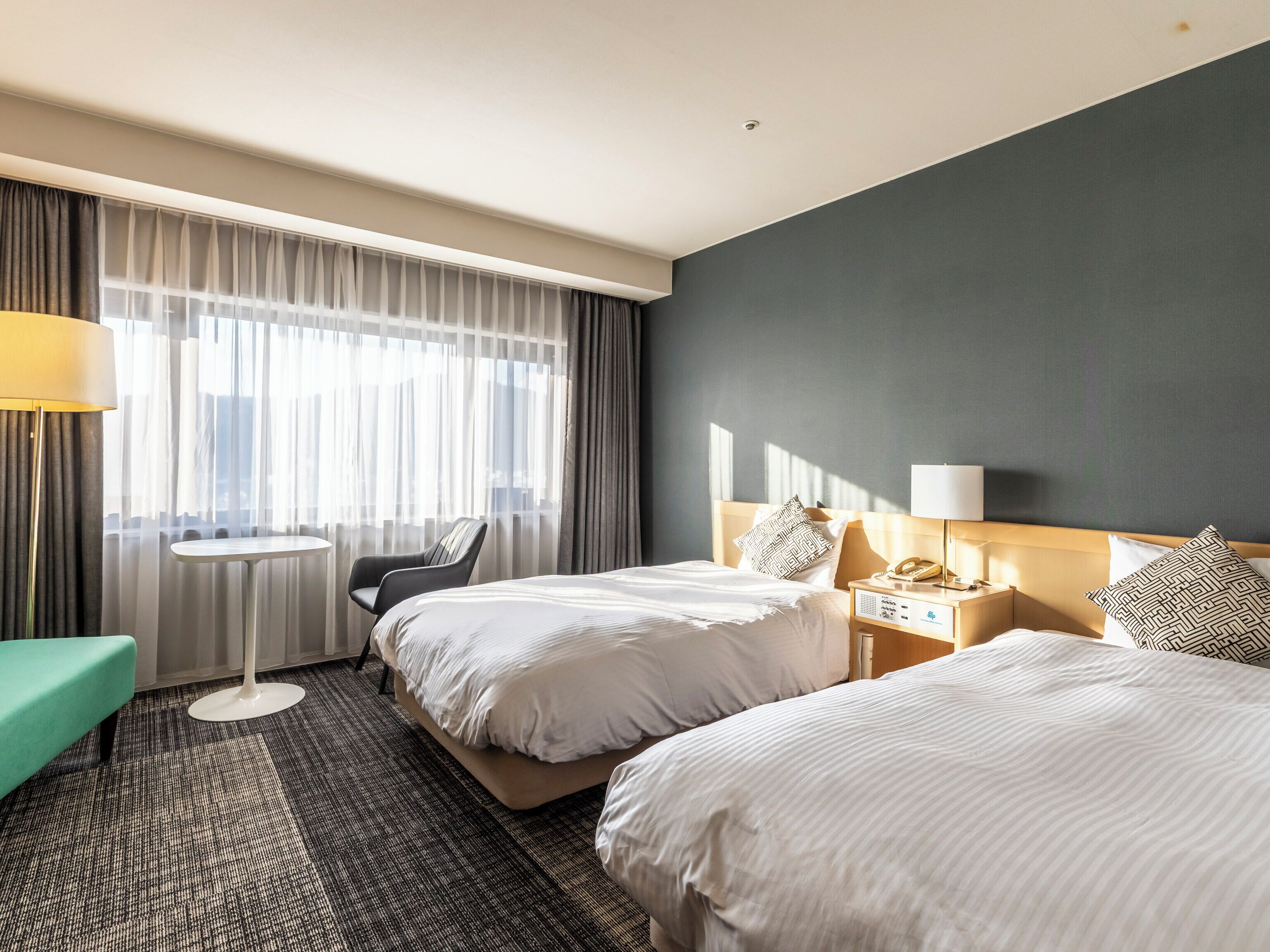 Habitación de hotel con dos camas, una mesita, una silla y vista a la ciudad a través de cortinas translúcidas.