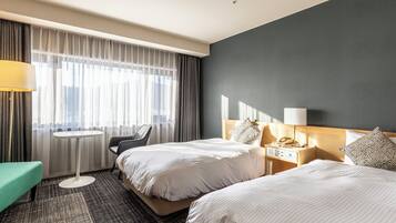 Habitación de hotel con dos camas, una mesita, una silla y vista a la ciudad a través de cortinas translúcidas.